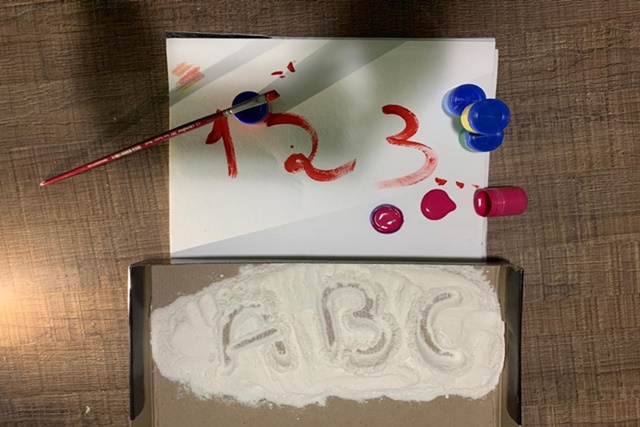 A imagem apresenta uma folha e um pedaço de papelão no chão. Na folha tem os números 1,2.3 pintados em vermelho. No papelão tem um pouco de areia e as letras a,b e c escritas.