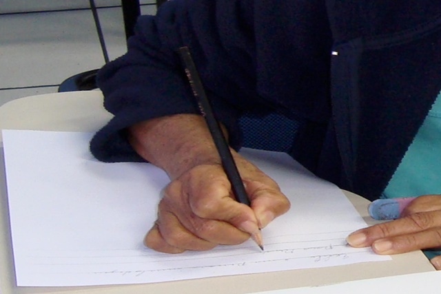 A foto tem uma mão escrevendo numa folha de papel com um lápis. O enquadramento da foto pega dos ombros para baixo. È uma pessoa branca e está vestida com uma camisa azul cuja as mangas vão até o ombro.